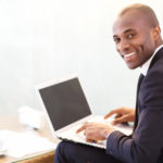 Businessman with laptop. Cheerful young African businessman typing something on laptop and looking over shoulder